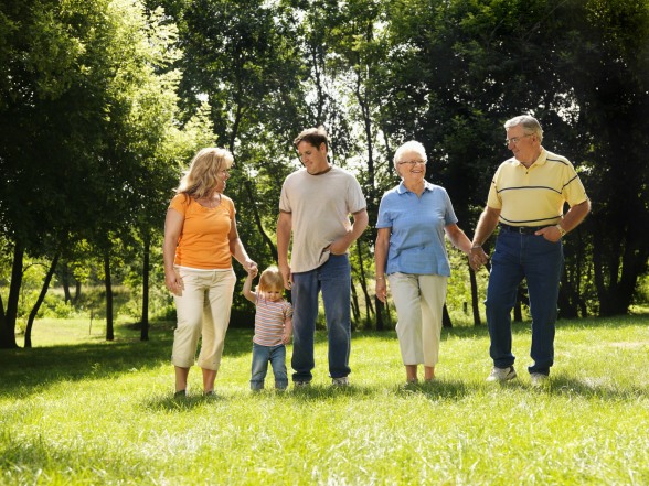 Three generation Caucasian family holding hands walking across grass in park smiling.